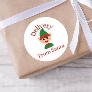 May include: Round white sticker with a red border. The sticker has a cartoon elf with a green hat and a red and white striped shirt. The text on the sticker says "Delivery from Santa".
