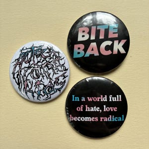 Op de afbeelding: Drie ronde buttons met grafische ontwerpen. Eén button heeft de tekst "BITE BACK" in wit en roze op een zwarte achtergrond. Een andere heeft de tekst "In a world full of hate, love becomes radical". De derde button heeft een witte achtergrond met een zwart-roze ontwerp.