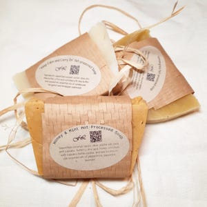 Puede incluir: Tres barras de jabón hecho a mano envueltas en papel marrón con etiquetas. Las etiquetas tienen un código QR y texto que describe los ingredientes y los aromas. Los jabones están etiquetados como "Keep Calm and Carry On", "Lemongrass" y "Honey & Mint".