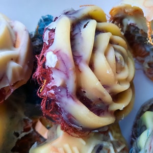 Könnte beinhalten: Nahaufnahme einer handgefertigten Seife in Form einer Rose. Die Seife ist hellgelb mit violetten und roten Wirbeln. Die Seife ist in ein rotes und weißes Tuch gewickelt.