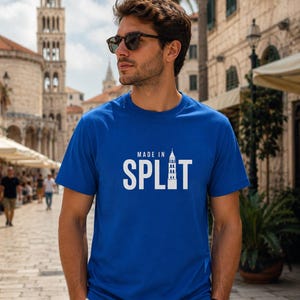 以下が含まれることがあります： 胸元に白い文字で「MADE IN SPLIT」とプリントされた鮮やかなブルーの半袖Tシャツ。文字の「I」の部分は、高い石造りの鐘楼のグラフィックイラストに置き換えられています。Tシャツはクルーネックの標準的なフィット感です。