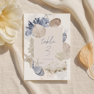 May include: A rectangular table number card with a beach theme. The card features watercolor illustrations of seashells, starfish, and coral in shades of blue, beige, and white. The text "table 3" is written in a script font, with the date "July 19th, 2027" below.