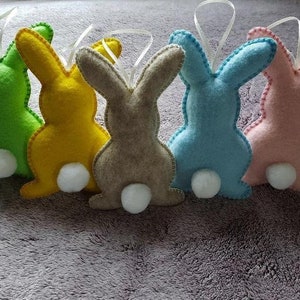 May include: Five felt bunny ornaments in green, yellow, brown, blue, and pink. Each bunny has a white pom-pom tail and a white ribbon for hanging. The edges are stitched with contrasting thread, perfect for Easter decorations.