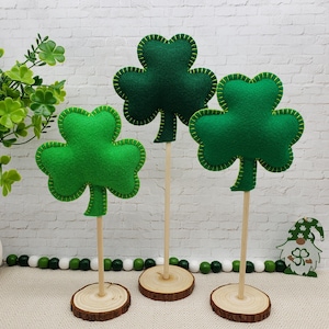May include: Three green felt shamrock decorations on wooden sticks with wooden bases. The shamrocks are outlined in green thread.