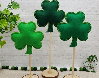 May include: Three green felt shamrock decorations on wooden sticks with wooden bases. The shamrocks are outlined in green thread.