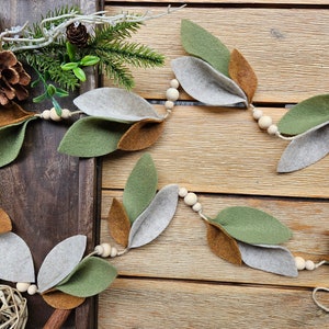 May include: A garland made of felt leaves in shades of green, brown, and gray, strung together with wooden beads. The garland is displayed on a wooden surface.