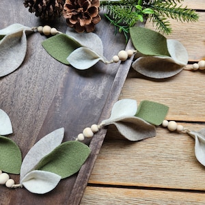May include: A decorative garland featuring felt leaves in shades of green and gray, strung together with wooden beads on a natural twine. The garland is displayed on a wooden tray, with a pinecone and greenery in the background.
