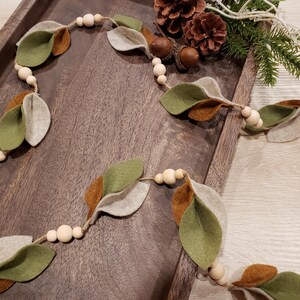 May include: A decorative garland featuring felt leaves in shades of green, brown, and gray, strung with wooden beads. The garland is arranged on a wooden tray, with pinecones and greenery in the background. A rustic, nature-inspired home decor item.