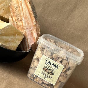 Puede incluir: Un recipiente de pl&aacute;stico transparente lleno de peque&ntilde;os trozos de arcilla de color tostado, etiquetado como Calaba Clay, 1 lb (454 g), junto a grandes rocas de arcilla cruda, estratificadas en tonos tostado y marr&oacute;n anaranjado.