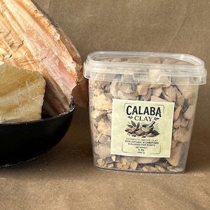 May include: A clear plastic container filled with small, irregular pieces of tan Calaba clay. The label reads Calaba Clay, Authentic Native Clay, 100% Natural & Unrefined, Sun-Dried for Purity, Net Weight 1 lb (454 g). A large, textured rock sits nearby.
