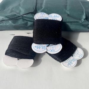 May include: Three spools of black thread with white labels, each marked with "91.44M". The spools are arranged on a white surface, with a crumpled green package in the background.