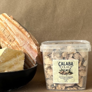 Puede incluir: Un recipiente de pl&aacute;stico transparente lleno de peque&ntilde;os trozos irregulares de arcilla Calaba, etiquetado con el texto 'CALABA CLAY, AUTHENTIC NATIVE CLAY, 100% NATURAL & UNREFINED, SUN-DRIED FOR PURITY, NET WEIGHT 1 lb (454 g)', junto a un cuenco negro de arcilla.