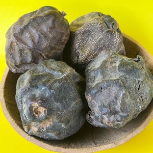 Puede incluir: Cuatro objetos oscuros de forma irregular, posiblemente frutos secos o nueces, están dispuestos en un cuenco de madera. Los objetos tienen una superficie rugosa y texturizada y un color marrón oscuro. El fondo es de color amarillo brillante.