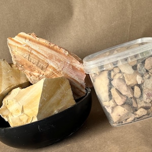 Puede incluir: Un cuenco negro que contiene tres piezas grandes y dentadas de roca sedimentaria de color amarillo claro y bronceado con texturas en capas visibles, junto a un recipiente de pl&aacute;stico transparente lleno de fragmentos irregulares de piedra similar.