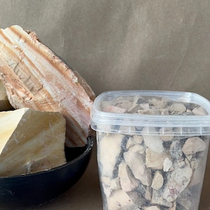 Puede incluir: Un recipiente de pl&aacute;stico transparente lleno de peque&ntilde;os trozos irregulares de arcilla o fragmentos de piedra de color beige, gris y blanco, junto a un cuenco negro que contiene dos grandes espec&iacute;menes de roca claros y estratificados con estr&iacute;as.