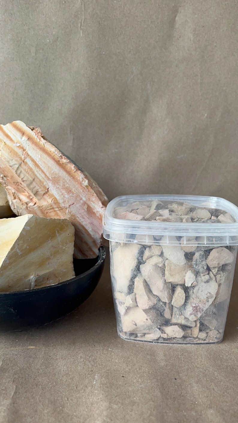 Puede incluir: Un recipiente de pl&aacute;stico transparente lleno de peque&ntilde;os trozos irregulares de arcilla o fragmentos de piedra de color beige, gris y blanco, junto a un cuenco negro que contiene dos grandes espec&iacute;menes de roca claros y estratificados con estr&iacute;as.