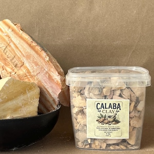 Puede incluir: Un recipiente de pl&aacute;stico transparente lleno de peque&ntilde;os trozos de arcilla Calaba de color tostado. La etiqueta dice: CALABA CLAY, AUTHENTIC NATIVE CLAY, 100% NATURAL & UNREFINED, SUN-DRIED FOR PURITY, NET WEIGHT 1 lb (454 g). Al lado hay un cuenco negro con grandes rocas de arcilla.