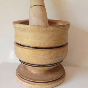 May include: A wooden mortar and pestle with a light brown finish and black accents. The mortar has a wide base and a rounded bowl. The pestle is cylindrical with a rounded top.
