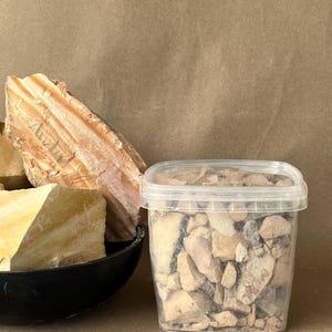 Puede incluir: Un recipiente rectangular de pl&aacute;stico transparente lleno de peque&ntilde;os fragmentos de roca irregulares de color beige y marr&oacute;n, junto a un cuenco negro que contiene grandes piedras minerales estratificadas de tonos crema y naranja con estr&iacute;as visibles.