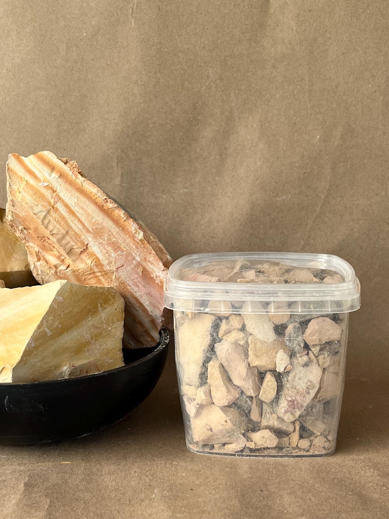Puede incluir: Un recipiente rectangular de pl&aacute;stico transparente lleno de peque&ntilde;os fragmentos de roca irregulares de color beige y marr&oacute;n, junto a un cuenco negro que contiene grandes piedras minerales estratificadas de tonos crema y naranja con estr&iacute;as visibles.