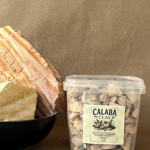 Puede incluir: Un recipiente de pl&aacute;stico transparente lleno de peque&ntilde;os trozos de arcilla de color tostado, etiquetado Calaba Clay, 1 lb (454 g), junto a un cuenco negro que contiene grandes rocas de arcilla cruda en tonos beige y naranja.