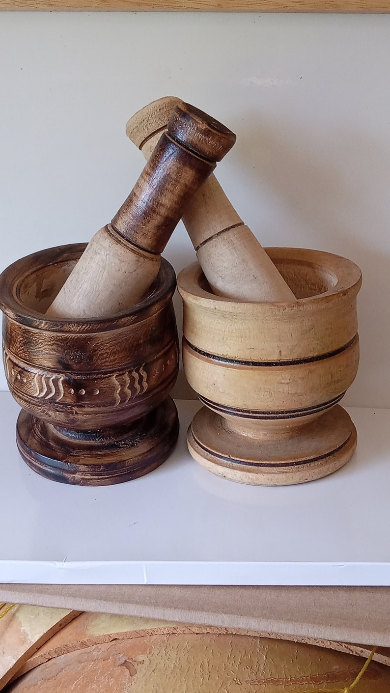 May include: Two wooden mortars and pestles, one with a dark brown finish and intricate carvings, the other with a light brown finish and simple bands. Both are made of natural wood and have a rustic, handcrafted look.