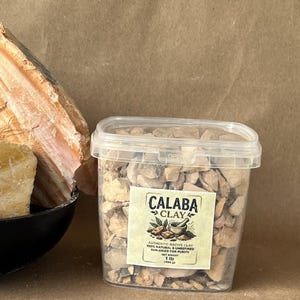 Puede incluir: Un recipiente de pl&aacute;stico transparente lleno de trozos de arcilla beige y marr&oacute;n. La etiqueta dice Calaba Clay, Authentic Native Clay, 100% Natural & Unrefined, Sun-Dried for Purity, Net Weight 1 lb (454 g). A la izquierda hay una roca grande, texturizada y de color beige.