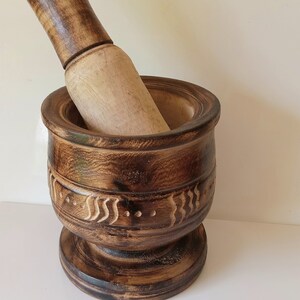 May include: A dark brown wooden mortar and pestle with a carved design. The mortar has a wide base and a deep bowl. The pestle is long and cylindrical.