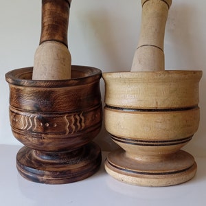 May include: Two wooden mortars and pestles, one brown with carved details and one light brown with black bands. Both are made of wood and have a round base.