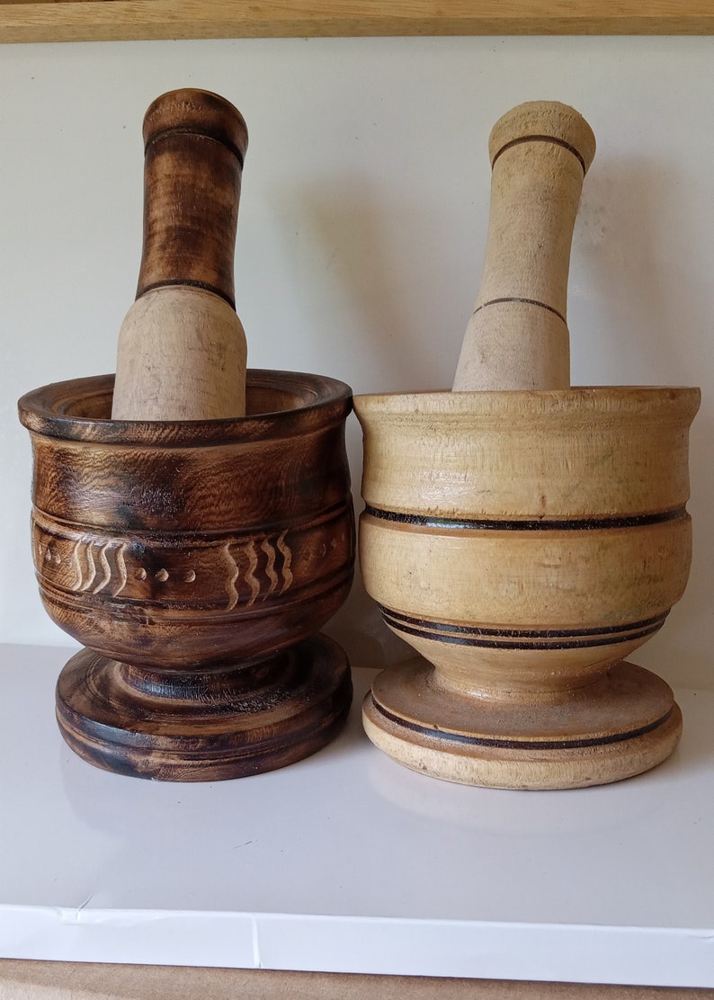 May include: Two wooden mortars and pestles, one brown with carved details and one light brown with black bands. Both are made of wood and have a round base.