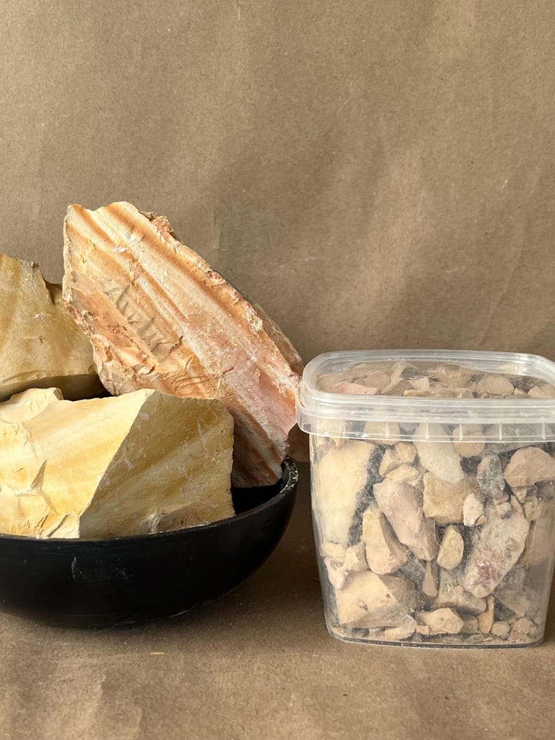 Puede incluir: Un cuenco negro que contiene trozos grandes y dentados de roca sedimentaria de color tostado y crema con capas horizontales visibles, junto a un recipiente de pl&aacute;stico transparente lleno de fragmentos m&aacute;s peque&ntilde;os del mismo material mineral.