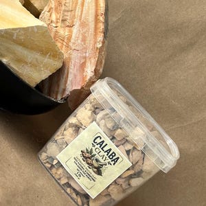 Puede incluir: Un recipiente de pl&aacute;stico transparente lleno de peque&ntilde;os trozos de arcilla de color beige, con una etiqueta que dice CALABA CLAY, 100% NATURAL & UNREFINED, SUN-DRIED FOR PURITY, 454 g. Al lado hay rocas de arcilla crudas, beige y anaranjadas.