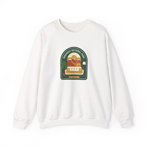 Könnte beinhalten: Weißes Sweatshirt mit einem grünen, orangefarbenen und gelben Aufdruck einer Berglandschaft und dem Text "INDOORSY NATIONAL PARK". Die Grafik zeigt ein Sofa, eine Lampe und eine Pflanze. Das Sweatshirt hat einen Rundhalsausschnitt und lange Ärmel.