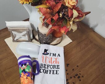 May include: A white kitchen towel with the text "I'm a Witch Before Coffee" in black lettering. A witch's hat with a black cat is above the text. The towel is folded and lying on a wooden table with coffee beans scattered around it. A purple travel mug with a Mickey Mouse Halloween design is next to the towel.