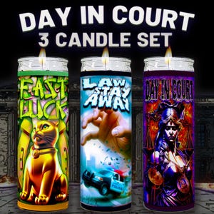 May include: A set of three glass jar candles with lit wicks. The candles have colourful labels with text and illustrations. The labels read "Fast Luck", "Law Stay Away", and "Day in Court". The candles are on a black background.