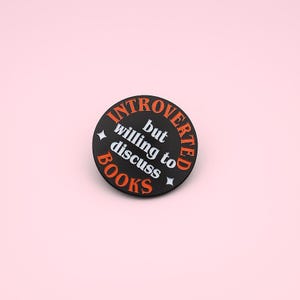 May include: A round, black pin with the words "INTROVERTED" and "BOOKS" in orange, and "but willing to discuss" in white. Two small white stars and a black border complete the design. The pin is set against a light pink background.