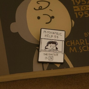 May include: A white and black enamel pin depicting a cartoon psychiatric help booth. The pin reads "PSYCHIATRIC HELP 5¢" and "THE DOCTOR IS IN". The pin is set against a book cover with a cartoon character.