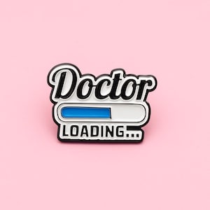 May include: A black and white enamel pin with the word "Doctor" in a script font above a loading bar design. The loading bar is white with a blue progress indicator. The text "LOADING..." is below the bar.