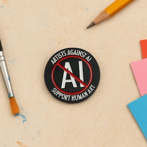 Puede incluir: Un pin circular negro y rojo con el texto "ARTISTS AGAINST AI" y "SUPPORT HUMAN ART". El centro presenta un símbolo "AI" blanco tachado con una línea roja. Un pincel y un lápiz están en el fondo.