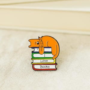 May include: Orange cat enamel pin resting on a stack of colorful books. The books are labeled "I Love Books" in white text. The pin is a cute accessory for book lovers.