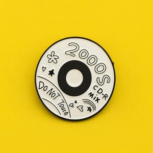 Puede incluir: Un pin de esmalte blanco y negro con forma de CD. El pin presenta el texto "2000s CD-R Mix" y "Do Not Touch", junto con pequeñas ilustraciones de una flor, corazones y estrellas. Sobre un fondo amarillo brillante.