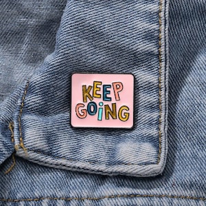 Puede incluir: Un pin de esmalte rosa con las palabras "KEEP GOING" en letras de bloque coloridas. El pin es rectangular con un borde negro y está sujeto a una chaqueta vaquera azul.