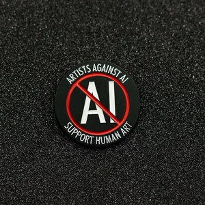 May include: A round, black pin with the text "ARTISTS AGAINST AI" and "SUPPORT HUMAN ART" in white lettering. The letters "AI" are crossed out with a red line. The pin is on a black, textured surface.