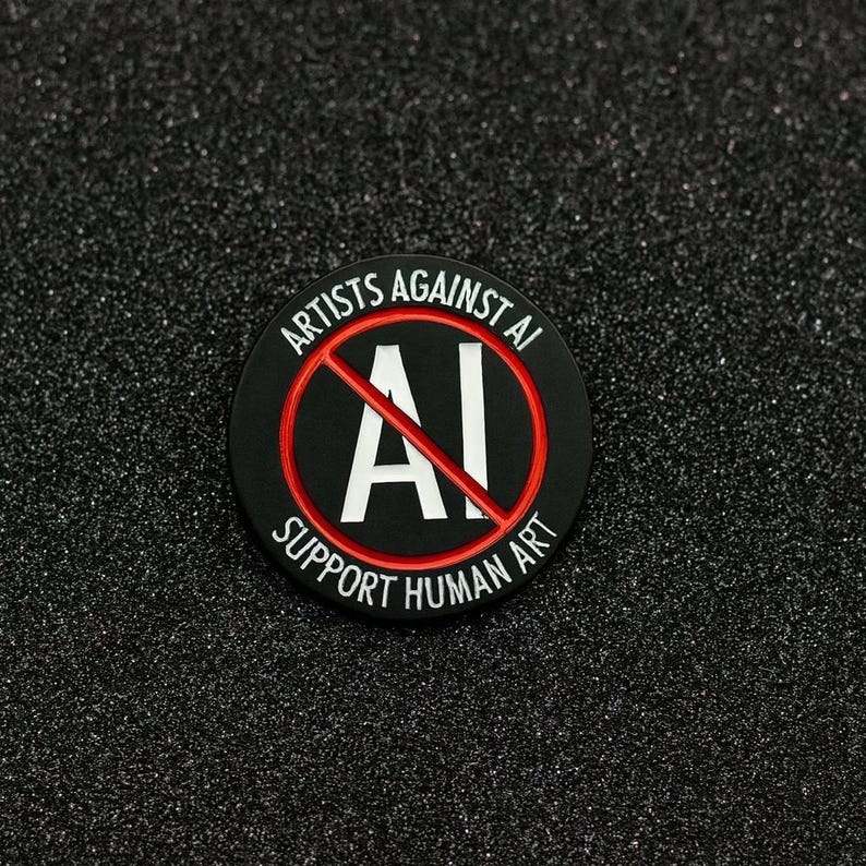 May include: A round, black pin with the text "ARTISTS AGAINST AI" and "SUPPORT HUMAN ART" in white lettering. The letters "AI" are crossed out with a red line. The pin is on a black, textured surface.