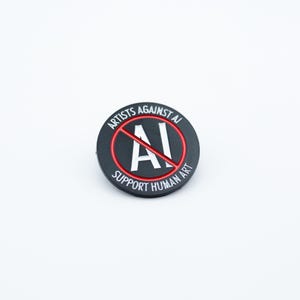 May include: A round, black pin with the text "ARTISTS AGAINST AI" and "SUPPORT HUMAN ART" in white lettering. A red circle with a line through the letters "AI" is in the center. The pin is on a white background.