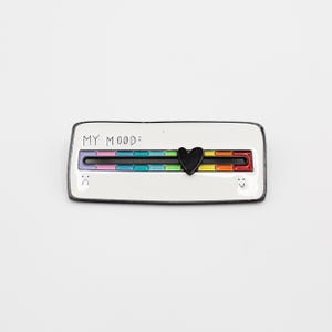 Puede incluir: Pin rectangular blanco con un control deslizante de estado de ánimo arcoíris y un indicador de corazón negro. El texto "MY MOOD:" está encima del control deslizante. Una cara triste está a la izquierda y una cara sonriente a la derecha.