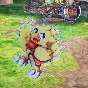 Könnte beinhalten: Eine gelbe und weiße Comic-Vogelfigur mit einer roten Tasche und einem Buch in den Krallen. Die Figur hält ein Buch und hat einen fröhlichen Ausdruck. Die Figur ist an einem silbernen Schlüsselanhänger befestigt.