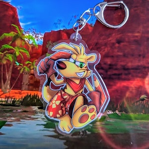 May include: A yellow and red cartoon character with a red bandana and a red and white patterned shorts. The character is holding a red and white object in its right hand. The character is on a clear acrylic keychain.