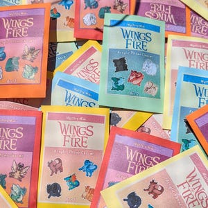 Puede incluir: Una colección de mini amuletos acrílicos para teléfono "Wings of Fire". El embalaje presenta el título en una fuente estilizada, con imágenes de los amuletos. El fondo es una mezcla de amarillo, naranja, rosa y azul.