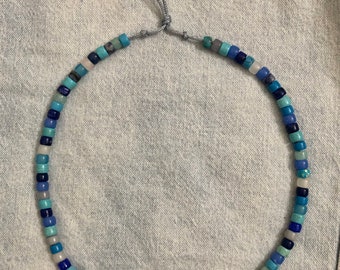 May include: A blue and white beaded necklace with a gray cord closure. The beads are various shades of blue and white, and the cord is tied in a knot at the back of the neck.